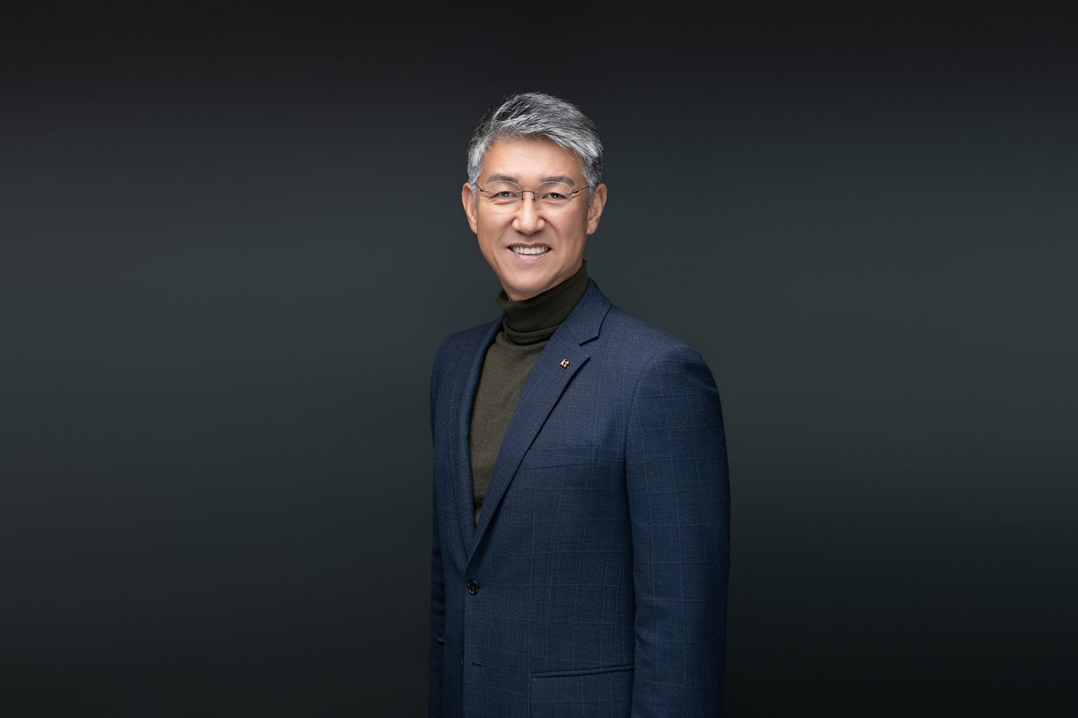 어두운 배경 앞에서 남색 재킷과 터틀넥을 입고 환하게 웃는 남성의 임원 프로필사진